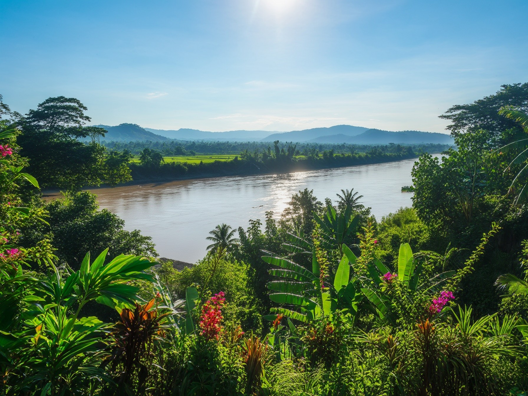 Scenic tropical landscape with lush greenery, vibrant flowers, river, and distant mountains under a clear blue sky.