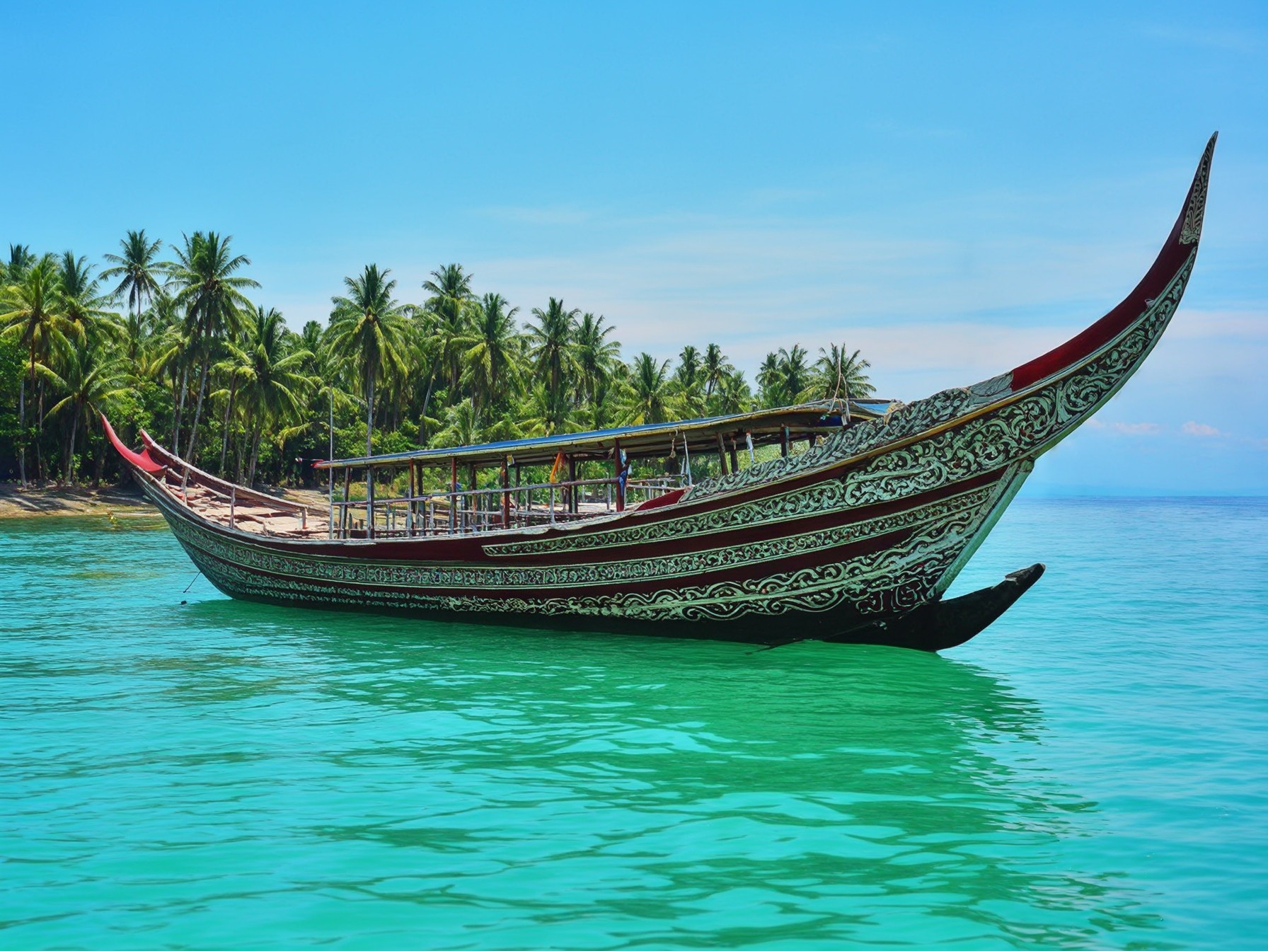 Traditional boat with ornate carvings on turquoise waters near tropical palm trees under clear blue sky.