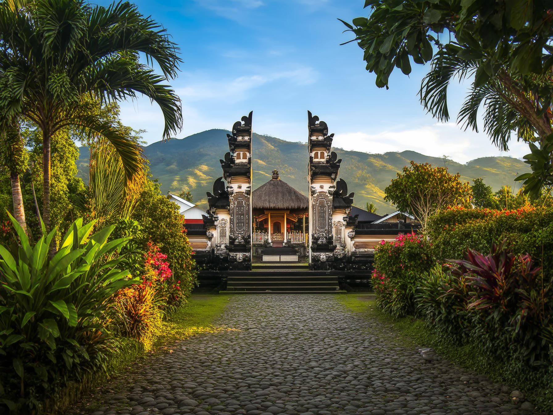 Traditional Bali temple gate surrounded by lush tropical greenery and mountains under a clear blue sky, Indonesia scenery.